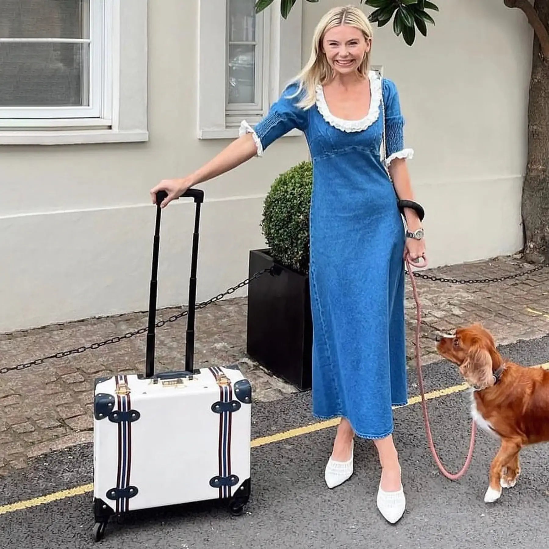 Woman in blue dress with white suitcase and dog, stylish travel look