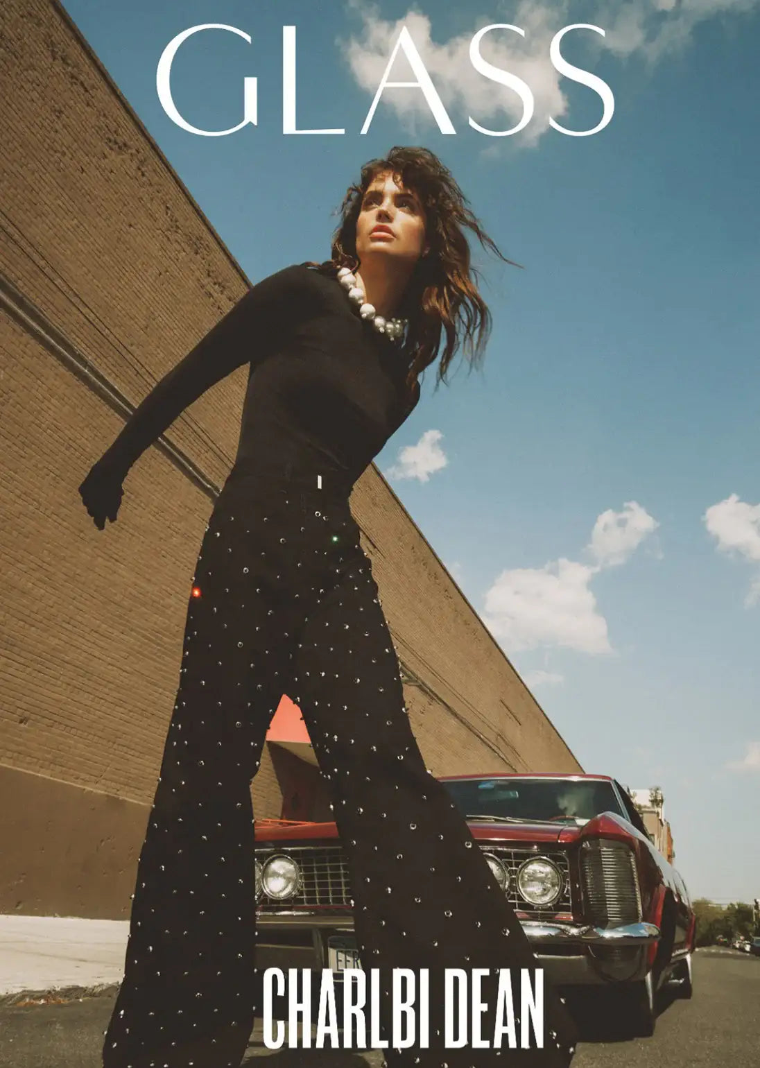 Charlbi Dean in black studded jumpsuit, pearl necklace, beside vintage car under blue sky