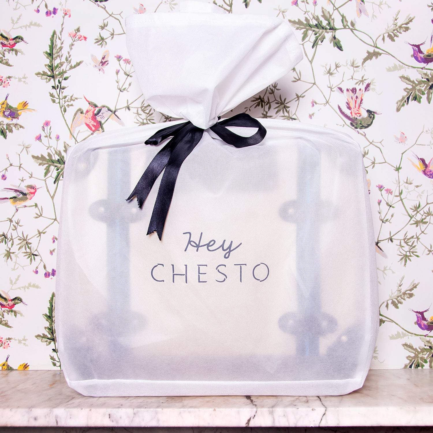 Hey CHESTO bag in sheer white wrap with black ribbon, set against floral bird wallpaper